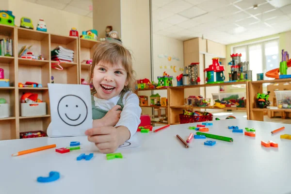 Little girl showing card with positive happy emotion during therapy session at kindergarten. Selective focus.