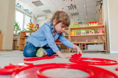 Anaokulunda ya da çocuk yuvasında yerde oturan kırmızı plastik oyuncaklarla oynayan küçük bir kız. Çocuk eğitimi konsepti.