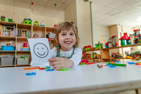 Little girl showing card with positive happy emotion during therapy session at kindergarten. Selective focus.