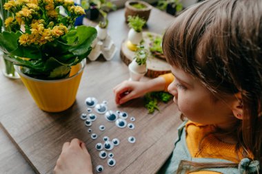Evdeki tahta masada yumurta kabuğunu oyuncak gözlerle süsleyen küçük bir kız. Bayramlık Paskalya dekorasyonu için yaratıcı bir fikir. Seçici odak.