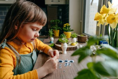 Evdeki tahta masada yumurta kabuğunu oyuncak gözlerle süsleyen küçük bir kız. Bayramlık Paskalya dekorasyonu için yaratıcı bir fikir. Seçici odak.