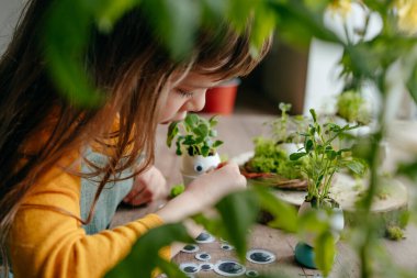 Küçük kız yumurta kabuklarını çimenle süslüyor. Evdeki ahşap masaya da oyuncak etiketler yapıştırıyor. Bayramlık Paskalya dekorasyonu için yaratıcı eğlenceli DIY fikri. Seçici odak.