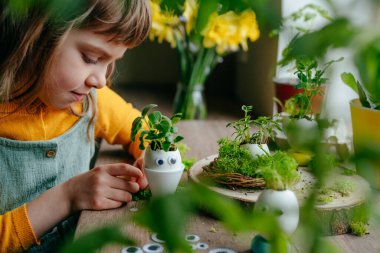 Küçük kız yumurta kabuklarını çimenle süslüyor. Evdeki ahşap masaya da oyuncak etiketler yapıştırıyor. Bayramlık Paskalya dekorasyonu için yaratıcı eğlenceli DIY fikri. Seçici odak.