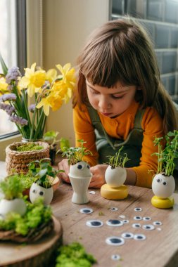 Küçük kız yumurta kabuklarını çimenle süslüyor. Evdeki ahşap masaya da oyuncak etiketler yapıştırıyor. Bayramlık Paskalya dekorasyonu için yaratıcı eğlenceli DIY fikri. Seçici odak.