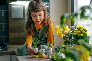 Küçük kız yumurta kabuklarını çimenle süslüyor. Evdeki ahşap masaya da oyuncak etiketler yapıştırıyor. Bayramlık Paskalya dekorasyonu için yaratıcı eğlenceli DIY fikri. Seçici odak.