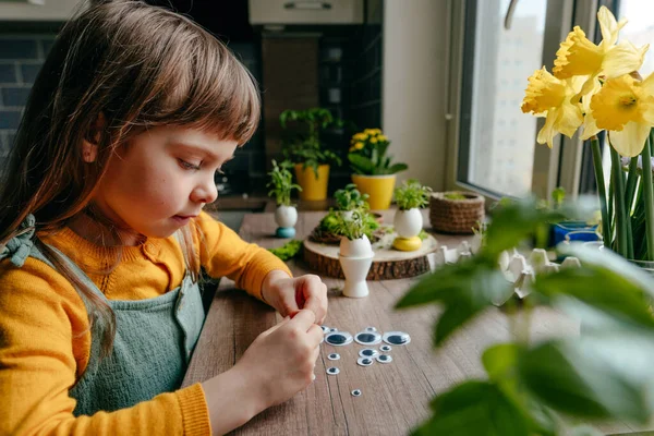 Evdeki tahta masada yumurta kabuğunu oyuncak gözlerle süsleyen küçük bir kız. Bayramlık Paskalya dekorasyonu için yaratıcı bir fikir. Seçici odak.