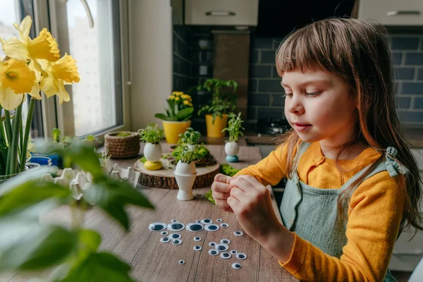 Evdeki ahşap masada yumurta kabuklarıyla çimleri süsleyen küçük bir kız. Bayramlık Paskalya dekorasyonu için yaratıcı eğlenceli DIY fikri. Seçici odak.