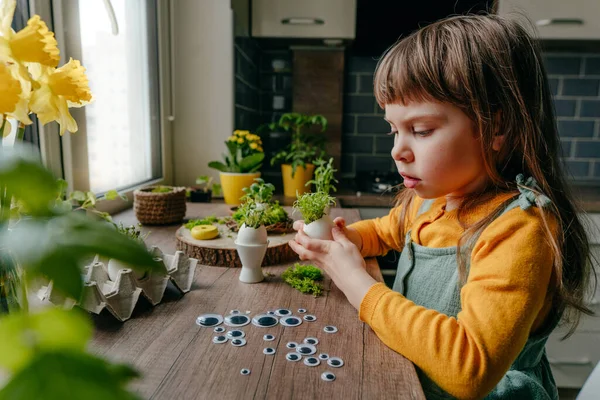 Evdeki ahşap masada yumurta kabuklarıyla çimleri süsleyen küçük bir kız. Bayramlık Paskalya dekorasyonu için yaratıcı eğlenceli DIY fikri. Seçici odak.