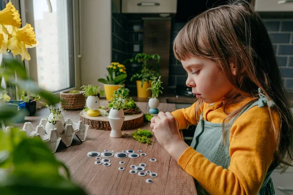 Evdeki ahşap masada yumurta kabuklarıyla çimleri süsleyen küçük bir kız. Bayramlık Paskalya dekorasyonu için yaratıcı eğlenceli DIY fikri. Seçici odak.