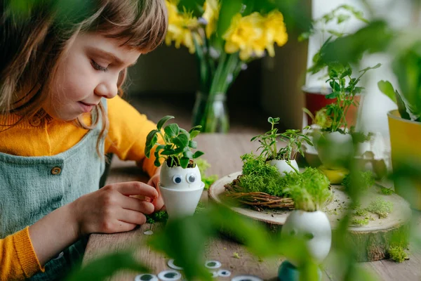 Küçük kız yumurta kabuklarını çimenle süslüyor. Evdeki ahşap masaya da oyuncak etiketler yapıştırıyor. Bayramlık Paskalya dekorasyonu için yaratıcı eğlenceli DIY fikri. Seçici odak.