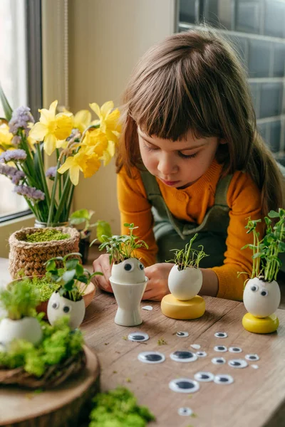 Küçük kız yumurta kabuklarını çimenle süslüyor. Evdeki ahşap masaya da oyuncak etiketler yapıştırıyor. Bayramlık Paskalya dekorasyonu için yaratıcı eğlenceli DIY fikri. Seçici odak.