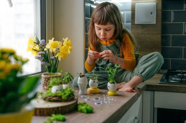 Küçük kız yumurta kabuklarını çimenle süslüyor. Evdeki ahşap masaya da oyuncak etiketler yapıştırıyor. Bayramlık Paskalya dekorasyonu için yaratıcı eğlenceli DIY fikri. Seçici odak.