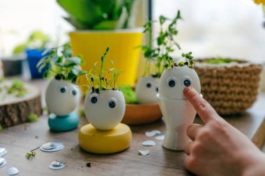 Küçük kız yumurta kabuklarını çimenle süslüyor. Evdeki ahşap masaya da oyuncak etiketler yapıştırıyor. Bayramlık Paskalya dekorasyonu için yaratıcı eğlenceli DIY fikri. Seçici odak.