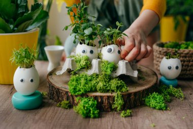 Evdeki ahşap masanın üzerinde oyuncak çıkartmalarla süslenmiş yumurta kabuklarıyla kompozisyon hazırlayan küçük kız. Bayramlık Paskalya dekorasyonu için yaratıcı eğlenceli DIY fikri. Seçici odak.