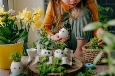 Evdeki ahşap masanın üzerinde oyuncak çıkartmalarla süslenmiş yumurta kabuklarıyla kompozisyon hazırlayan küçük kız. Bayramlık Paskalya dekorasyonu için yaratıcı eğlenceli DIY fikri. Seçici odak.
