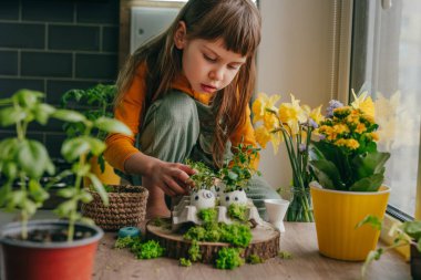 Evdeki ahşap masanın üzerinde oyuncak çıkartmalarla süslenmiş yumurta kabuklarıyla kompozisyon hazırlayan küçük kız. Bayramlık Paskalya dekorasyonu için yaratıcı eğlenceli DIY fikri. Seçici odak.