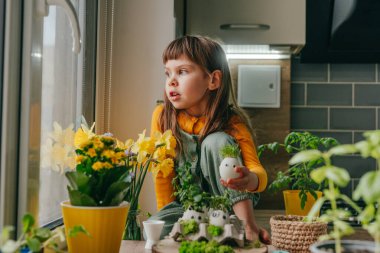 Evdeki ahşap masanın üzerinde oyuncak çıkartmalarla süslenmiş yumurta kabuklarıyla kompozisyon hazırlayan küçük kız. Bayramlık Paskalya dekorasyonu için yaratıcı eğlenceli DIY fikri. Seçici odak.