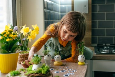 Küçük kız yumurta kabuklarını çimenle süslüyor. Evdeki ahşap masaya da oyuncak etiketler yapıştırıyor. Bayramlık Paskalya dekorasyonu için yaratıcı eğlenceli DIY fikri. Seçici odak.
