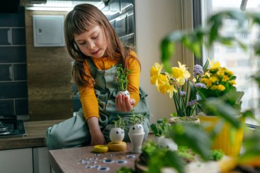Küçük kız yumurta kabuklarını çimenle süslüyor. Evdeki ahşap masaya da oyuncak etiketler yapıştırıyor. Bayramlık Paskalya dekorasyonu için yaratıcı eğlenceli DIY fikri. Seçici odak.