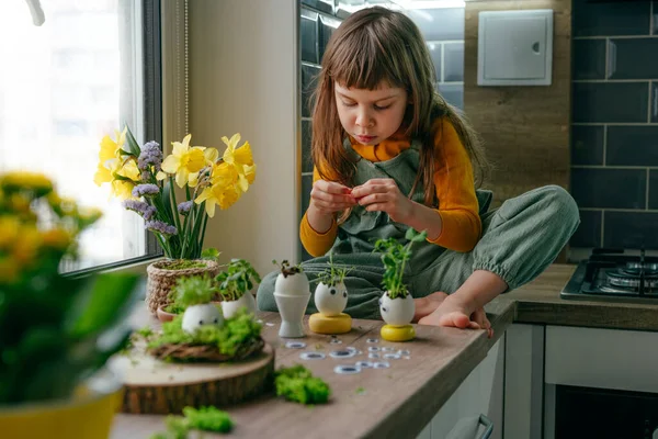 Küçük kız yumurta kabuklarını çimenle süslüyor. Evdeki ahşap masaya da oyuncak etiketler yapıştırıyor. Bayramlık Paskalya dekorasyonu için yaratıcı eğlenceli DIY fikri. Seçici odak.