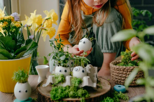 Evdeki ahşap masanın üzerinde oyuncak çıkartmalarla süslenmiş yumurta kabuklarıyla kompozisyon hazırlayan küçük kız. Bayramlık Paskalya dekorasyonu için yaratıcı eğlenceli DIY fikri. Seçici odak.