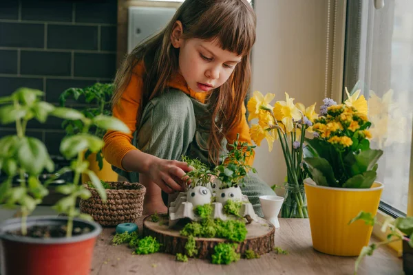 Evdeki ahşap masanın üzerinde oyuncak çıkartmalarla süslenmiş yumurta kabuklarıyla kompozisyon hazırlayan küçük kız. Bayramlık Paskalya dekorasyonu için yaratıcı eğlenceli DIY fikri. Seçici odak.