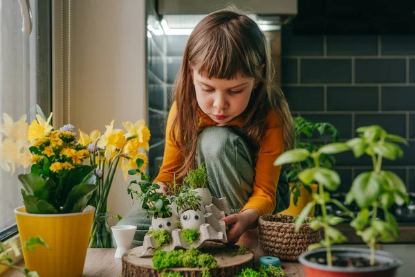 Evdeki ahşap masanın üzerinde oyuncak çıkartmalarla süslenmiş yumurta kabuklarıyla kompozisyon hazırlayan küçük kız. Bayramlık Paskalya dekorasyonu için yaratıcı eğlenceli DIY fikri. Seçici odak.