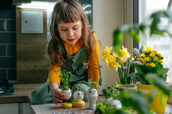 Küçük kız yumurta kabuklarını çimenle süslüyor. Evdeki ahşap masaya da oyuncak etiketler yapıştırıyor. Bayramlık Paskalya dekorasyonu için yaratıcı eğlenceli DIY fikri. Seçici odak.