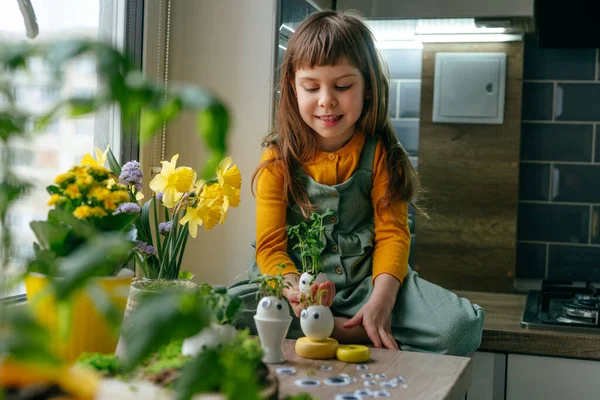 Küçük kız yumurta kabuklarını çimenle süslüyor. Evdeki ahşap masaya da oyuncak etiketler yapıştırıyor. Bayramlık Paskalya dekorasyonu için yaratıcı eğlenceli DIY fikri. Seçici odak.