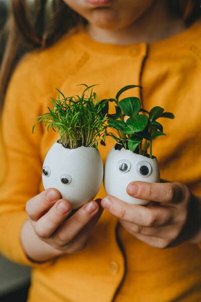 Little girl holding eggshells with cucumber and carrot sprouts with toy stickers eyes. Creative fun DIY idea for festive Easter decoration. Selective focus.