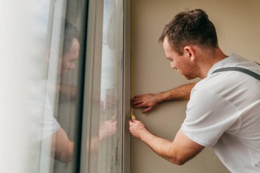 Genç adam, balkonda su geçirmez silikon kalıntı kullanarak pencereyi mühürlemeden önce tulum giyip bant yapıştırıyor..