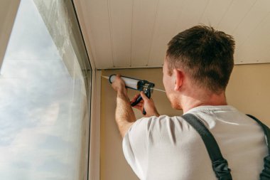 Balkonda su geçirmez silikon caulk kullanarak pencere ile süsleme arasındaki çatlakları kapatan tulum giyen genç bir adam..