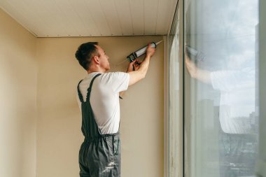 Young man wearing overalls sealing cracks between window and trim using waterproof silicone caulk on the balcony.