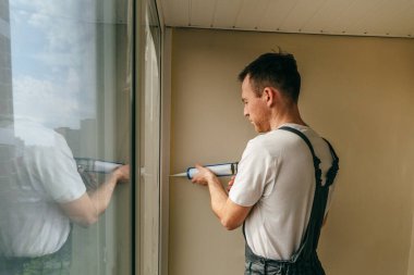 Balkonda su geçirmez silikon caulk kullanarak pencere ile süsleme arasındaki çatlakları kapatan tulum giyen genç bir adam..