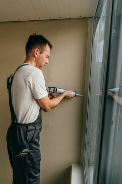 Balkonda su geçirmez silikon caulk kullanarak pencere ile süsleme arasındaki çatlakları kapatan tulum giyen genç bir adam..