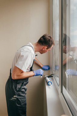 Genç adam tulum giyip balkonda su geçirmez silikon kazan kullanarak çatlakları kapatıyor. Lastik spatula kullanarak dolguyu çıkarıyorum. Seçici odak.
