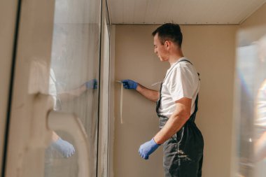 Genç adam tulum giyip balkonda su geçirmez silikon kazan kullanarak çatlakları kapatıyor. Bağlama bandını çıkarıyorum. Seçici odak.