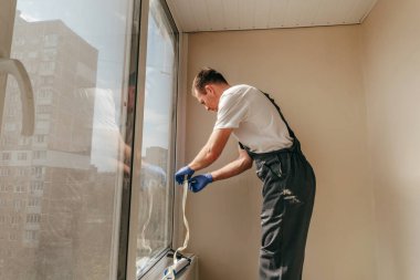 Genç adam tulum giyip balkonda su geçirmez silikon kazan kullanarak çatlakları kapatıyor. Bağlama bandını çıkarıyorum. Seçici odak.