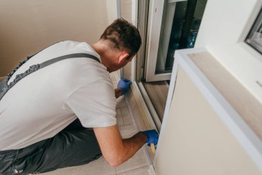 Genç adam, balkondaki su geçirmez silikon kalkanı kullanarak kapıyı mühürlemeden önce tulum giyip bant yapıştırıyordu. Seçici odak.