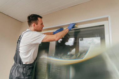 Balkona cam geçirmez silikon kalkanı yapıştırmadan önce tulum giymiş genç bir adam montaj bandı takıyordu. Seçici odak.