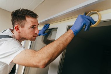 Balkona cam geçirmez silikon kalkanı yapıştırmadan önce tulum giymiş genç bir adam montaj bandı takıyordu. Seçici odak.