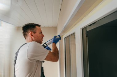 Balkonda su geçirmez silikon caulk kullanarak pencere ile süsleme arasındaki çatlakları kapatan tulum giyen genç bir adam..