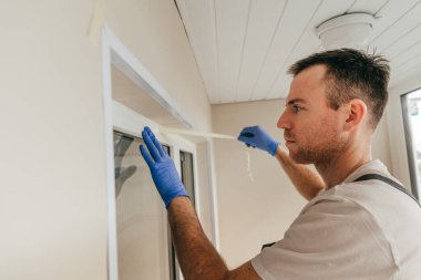 Genç adam tulum giyip balkonda su geçirmez silikon kazan kullanarak çatlakları kapatıyor. Bağlama bandını çıkarıyorum. Seçici odak.