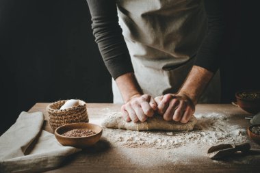 Ahşap masada hamur yoğuran tanınmaz genç adam. Erkeklerin elleri arka planda ekmek yapıyor. Seçici odak.