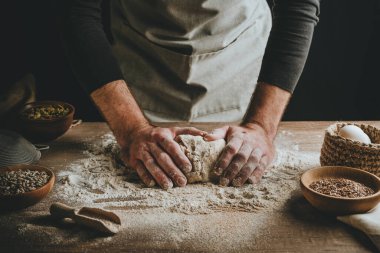 Unrecognizable young man kneading dough on wooden table. Males hands making bread on dark background. Selective focus.