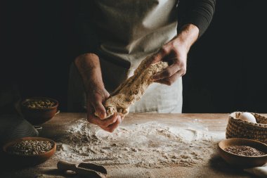 Ahşap masada hamur yoğuran tanınmaz genç adam. Erkeklerin elleri arka planda ekmek yapıyor. Seçici odak.
