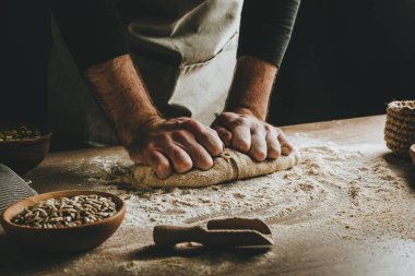 Ahşap masada hamur yoğuran tanınmaz genç adam. Erkeklerin elleri arka planda ekmek yapıyor. Seçici odak.