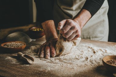 Unrecognizable young man kneading dough on wooden table. Males hands making bread on dark background. Selective focus.