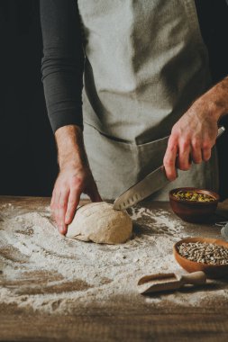 Unrecognizable young man making cuts with knife on uncooked fresh bread on wooden table on dark background. Selective focus.