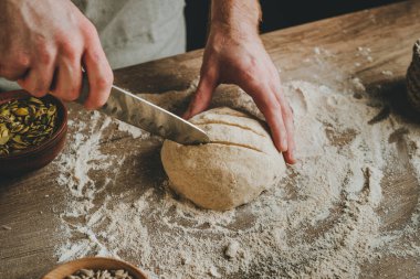 Tanımlanamayan genç adam, karanlık arka plandaki ahşap masada pişmemiş taze ekmek üzerinde bıçakla kesikler yapıyor. Seçici odak.